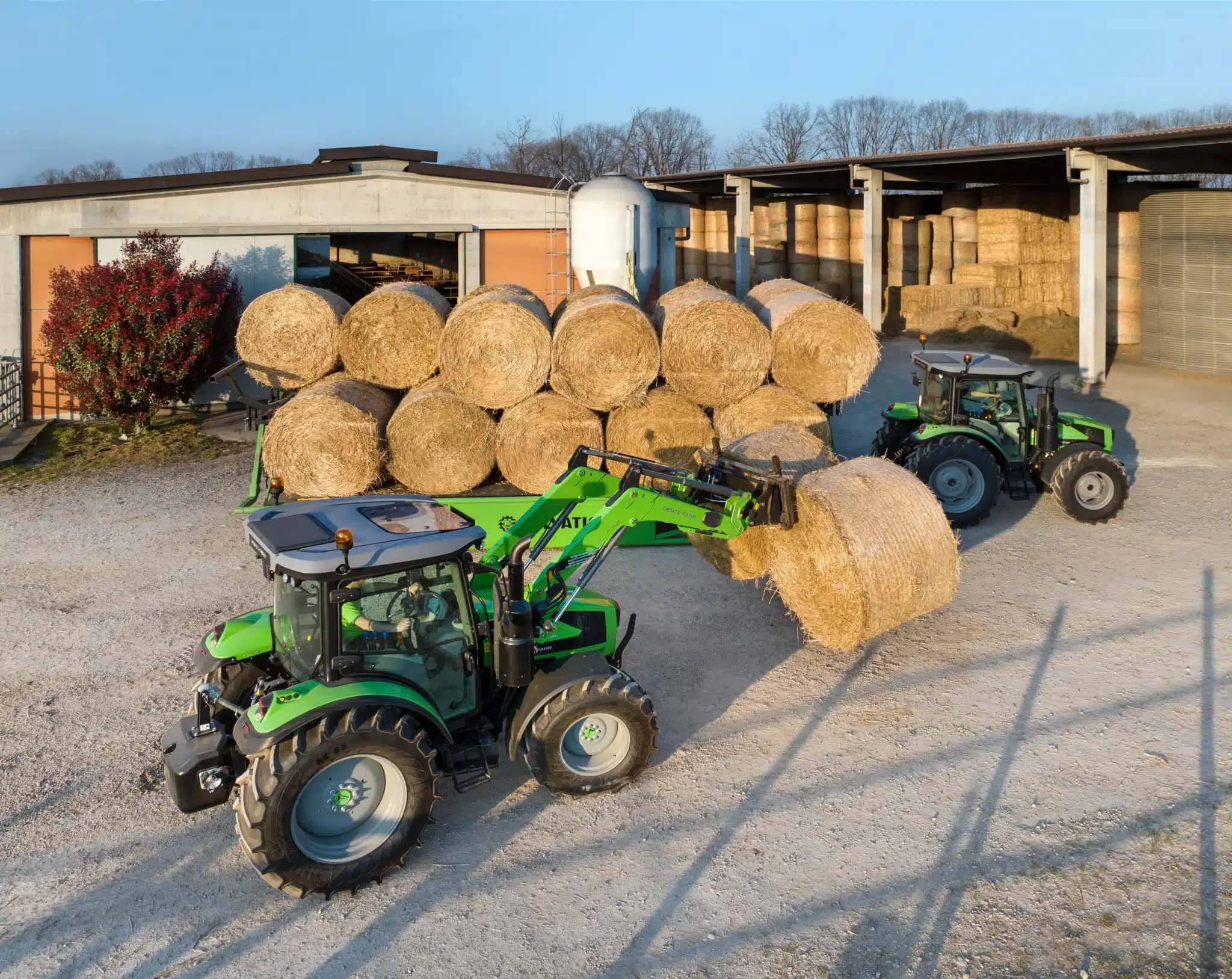 Grøn DEUTZ-FAHR traktor med frontlæsser der løfter en rund halmballe foran en gårds bygninger med stablede halmballer.
