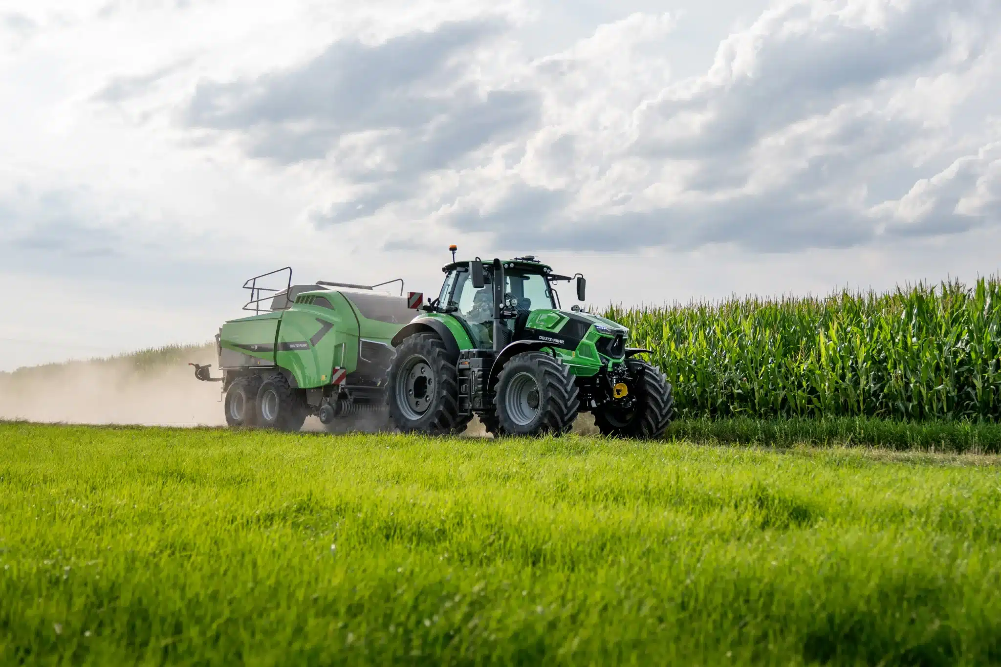 DEUTZ-FAHR traktor trækker en stor rundballepresser hen over marken langs en majsmark på en støvet sommerdag.