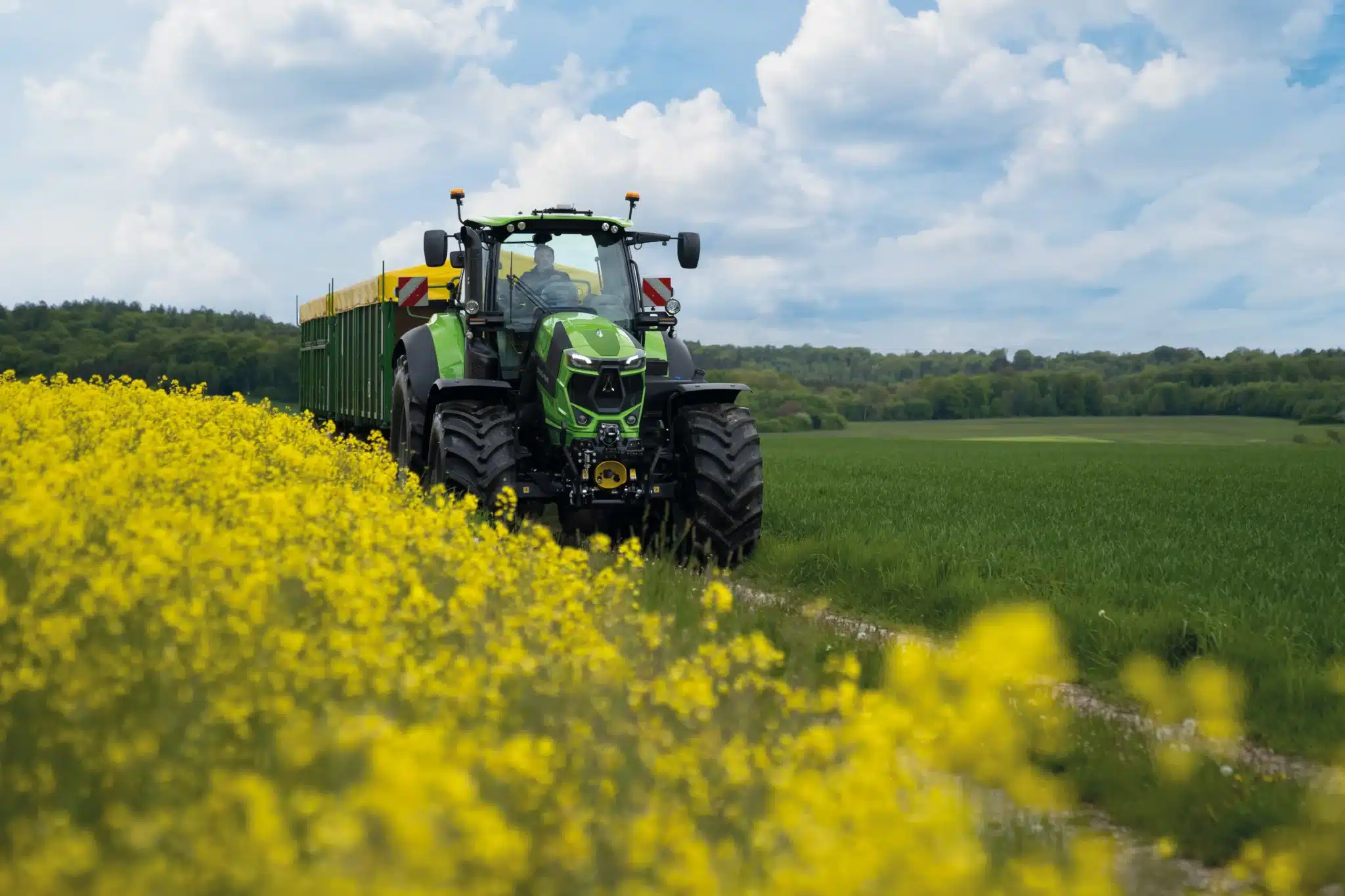 DEUTZ-FAHR traktor med tipvogn kører langs en markvej omgivet af gule rapsblomster og grønne marker.