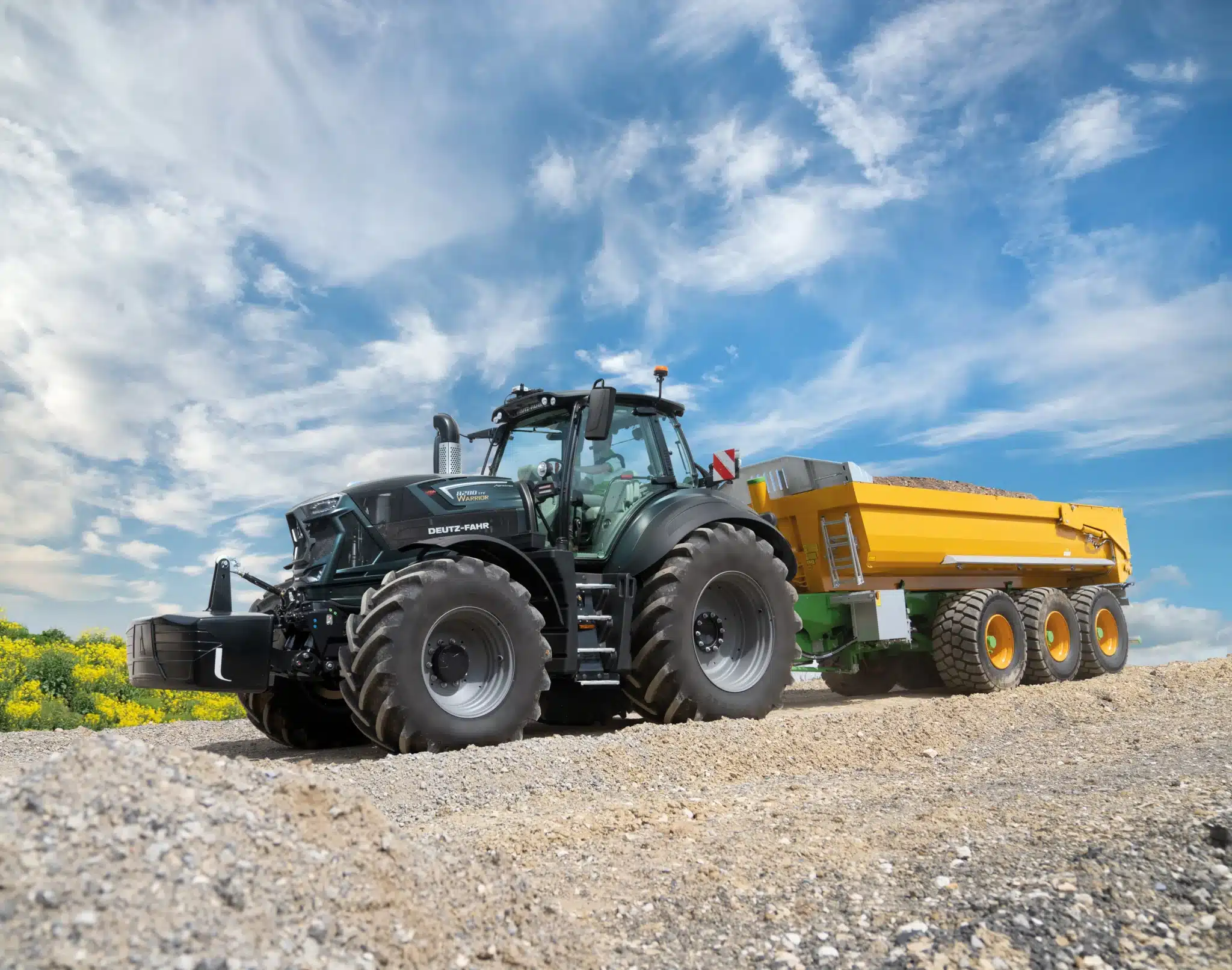 Sort DEUTZ-FAHR Warrior traktor med stor gul tipvogn kører på grusvej under blå himmel.